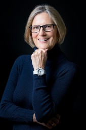 Black background with woman with spectacles and short bobbed fair hair wearing a black polo neck jumper